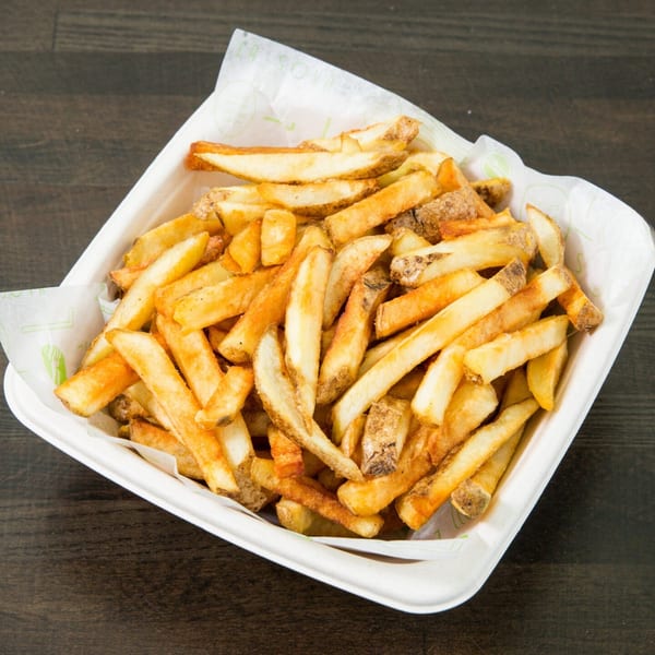 Golden-brown, skin-on french fries piled high in a white paper container on a dark wooden surface.